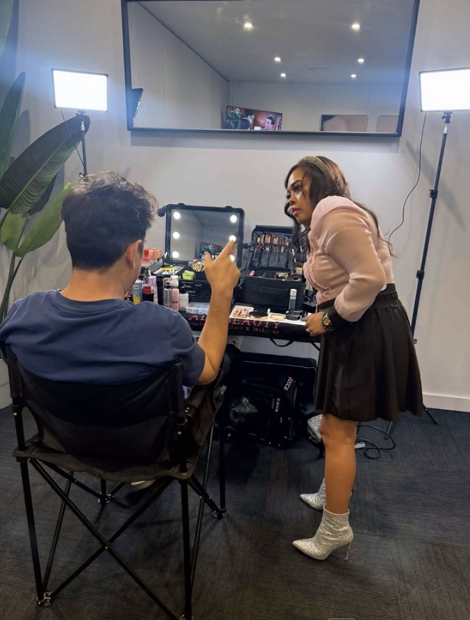 Makeup application backstage at an event, studio lighting rig either side